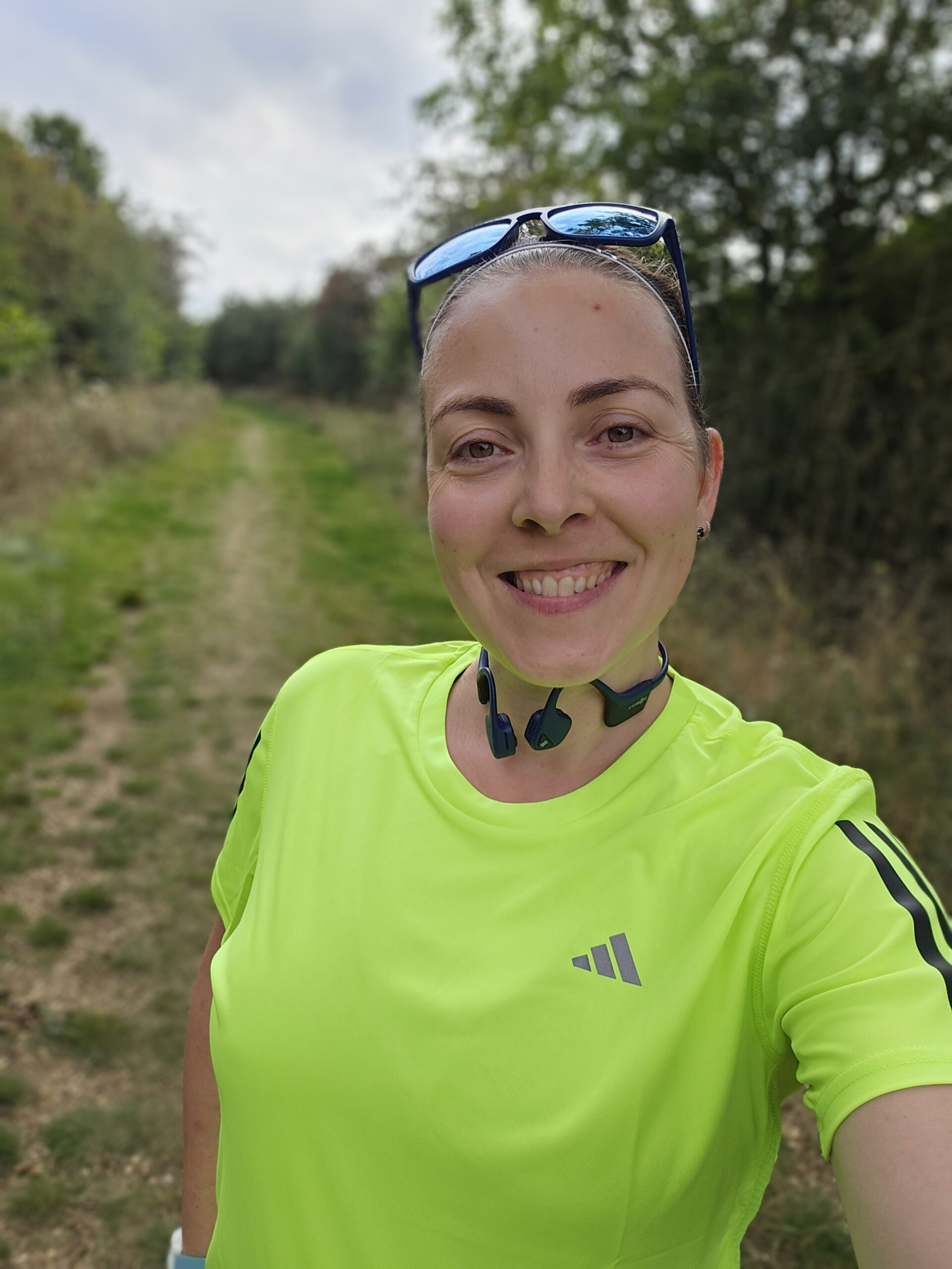 woman in yellow running top smiles at the camera, with a green grassy trail behind her