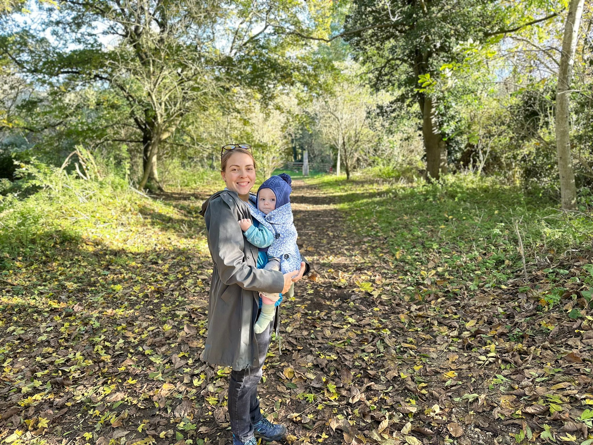 woman in green coat is carrying a baby in a sling. she is smiling at the camera and the baby is wearing a blue woolly hat. They are on a muddy path through the woods with trees behind them.