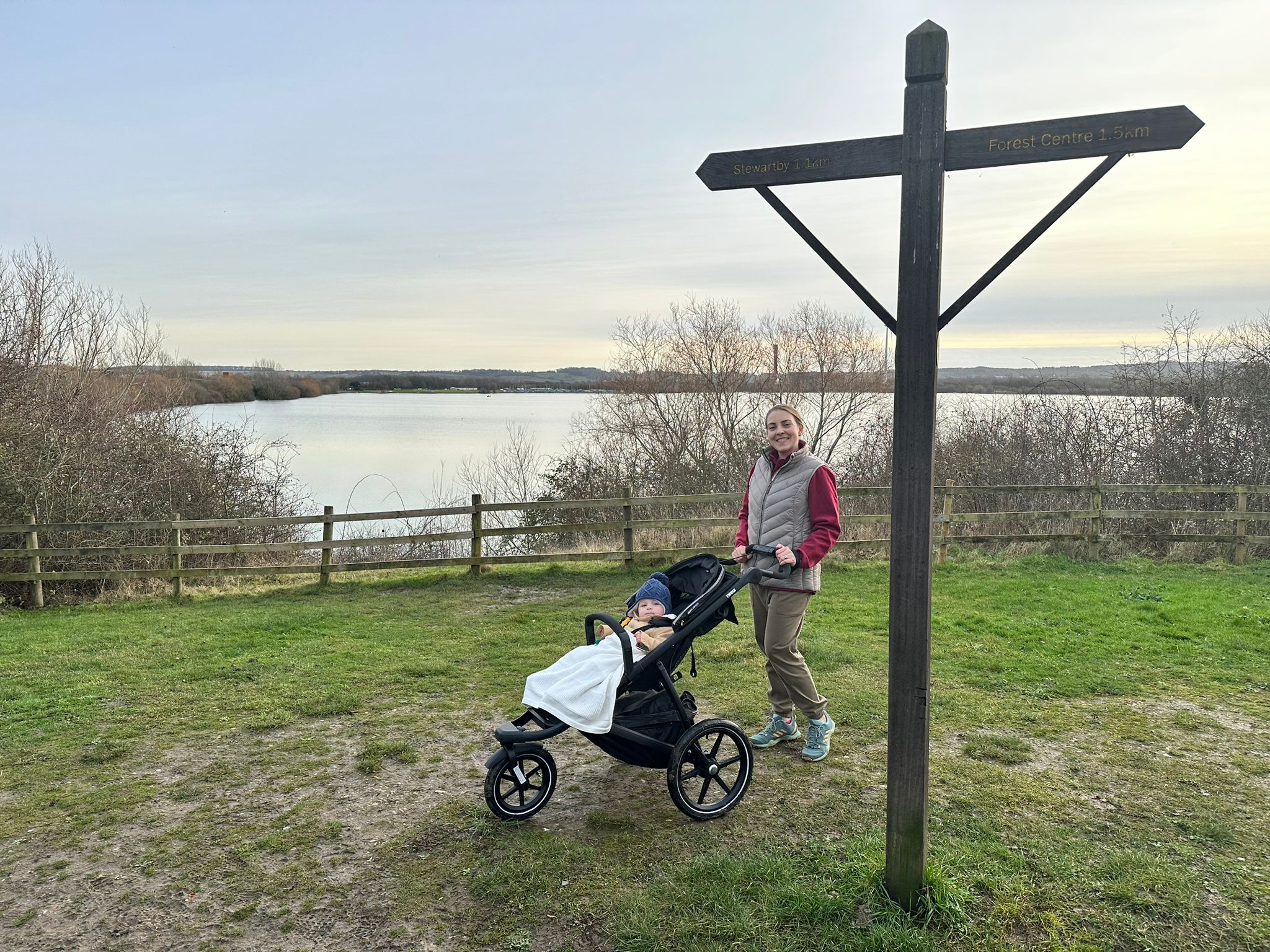 Woman with a running buggy stands on the grass by a wooden sign, with a lake in the background