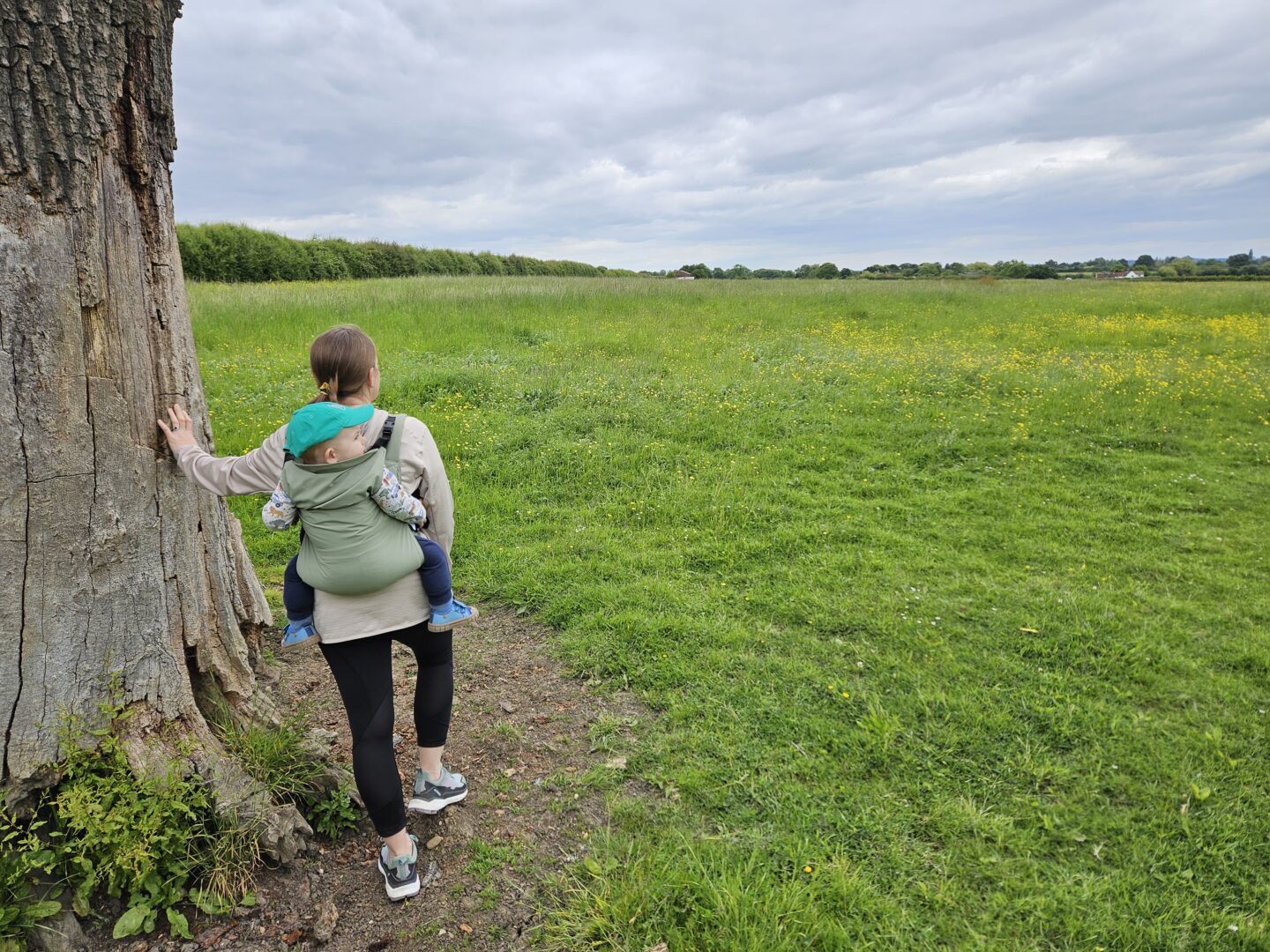 woman carrying baby on her back in carrier. she is leaning on a tree in the middle of a green field.