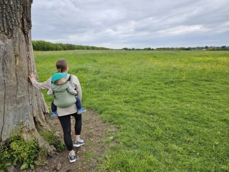 woman carrying baby on her back in carrier. she is leaning on a tree in the middle of a green field.