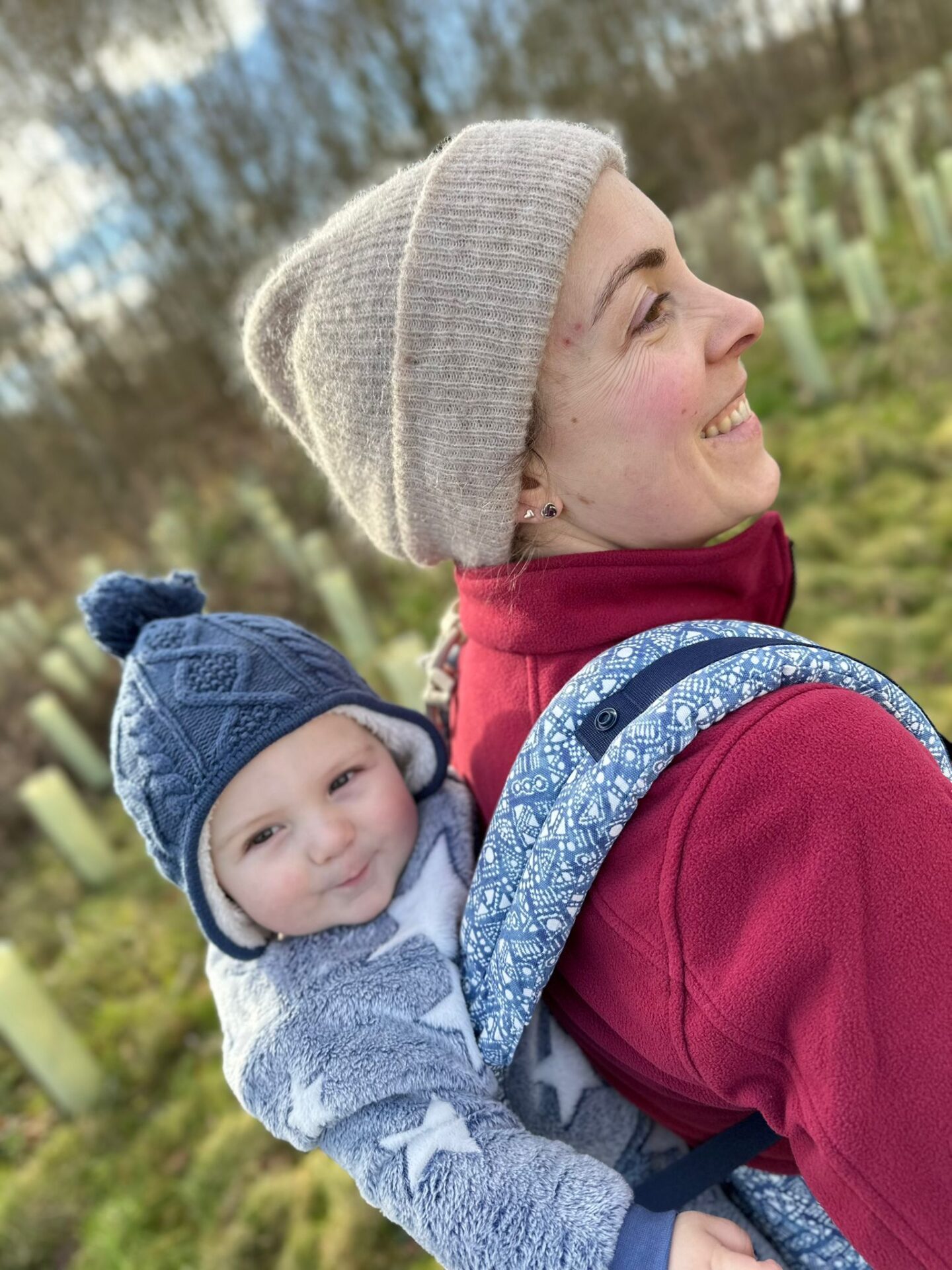 smiling woman in a beanie hat is carrying a toddler in a carrier on her back. 