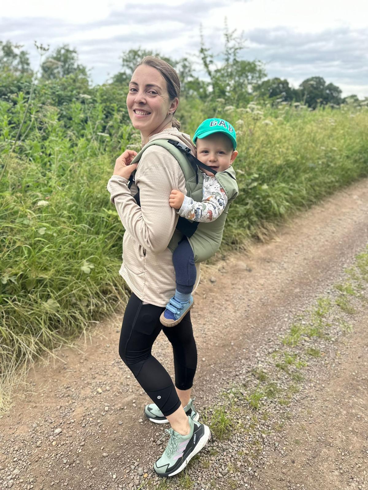 woman carrying toddler in a carrier on her back. she is standing on a gravel path with fields behind them