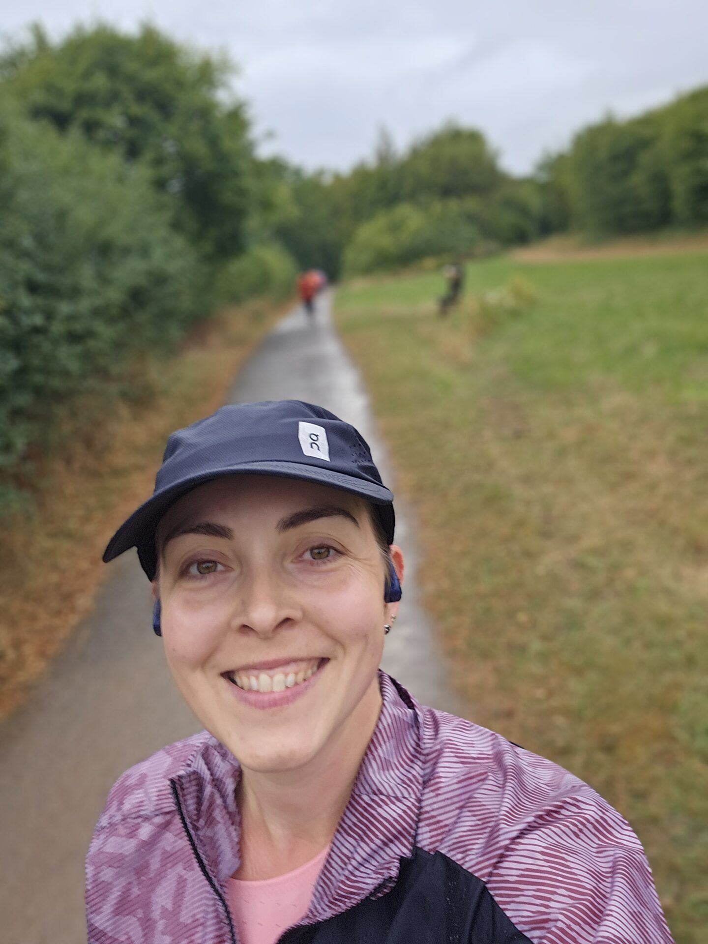 Selfie of a woman in a purple running jacket and black cap, standing on a footpath