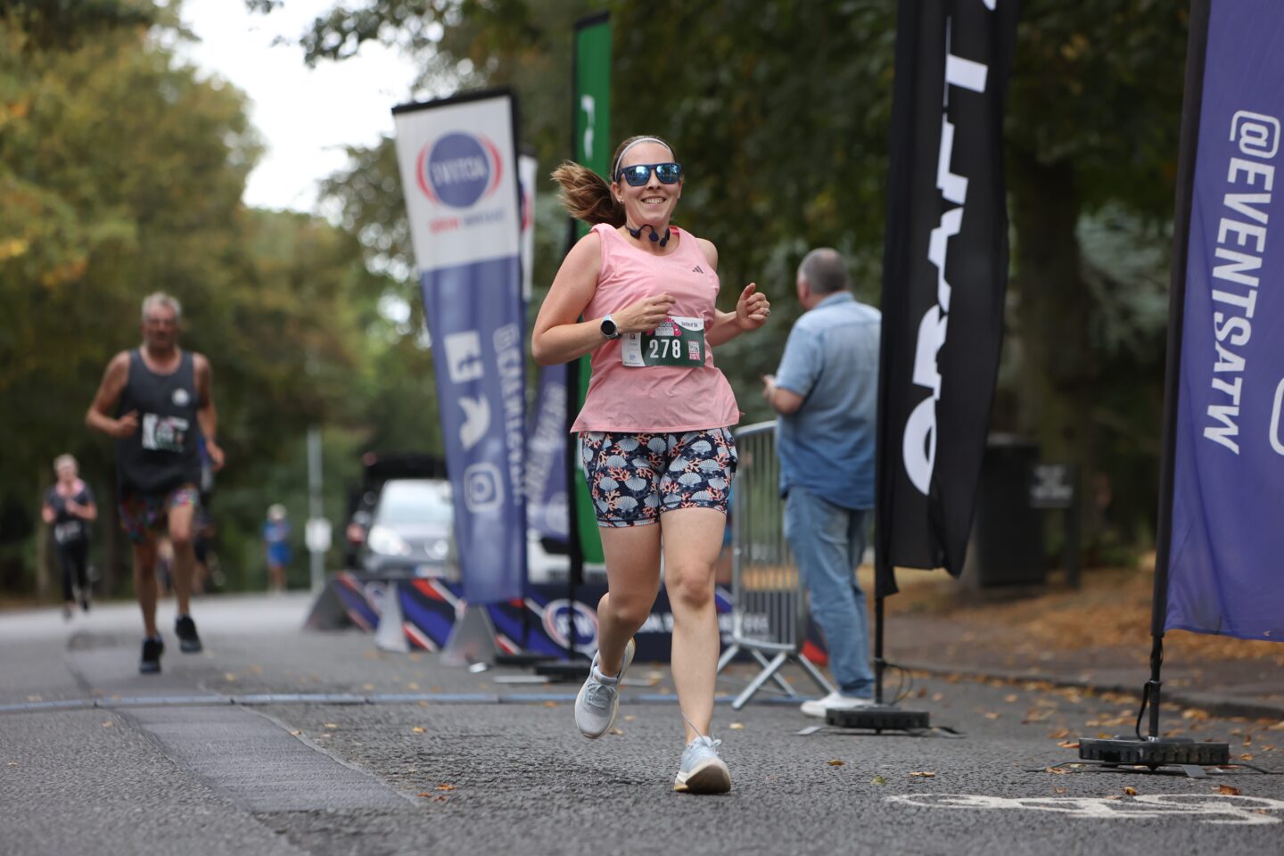 Woman in a pink tank top and shorts is smiling at the camera as she runs along the road