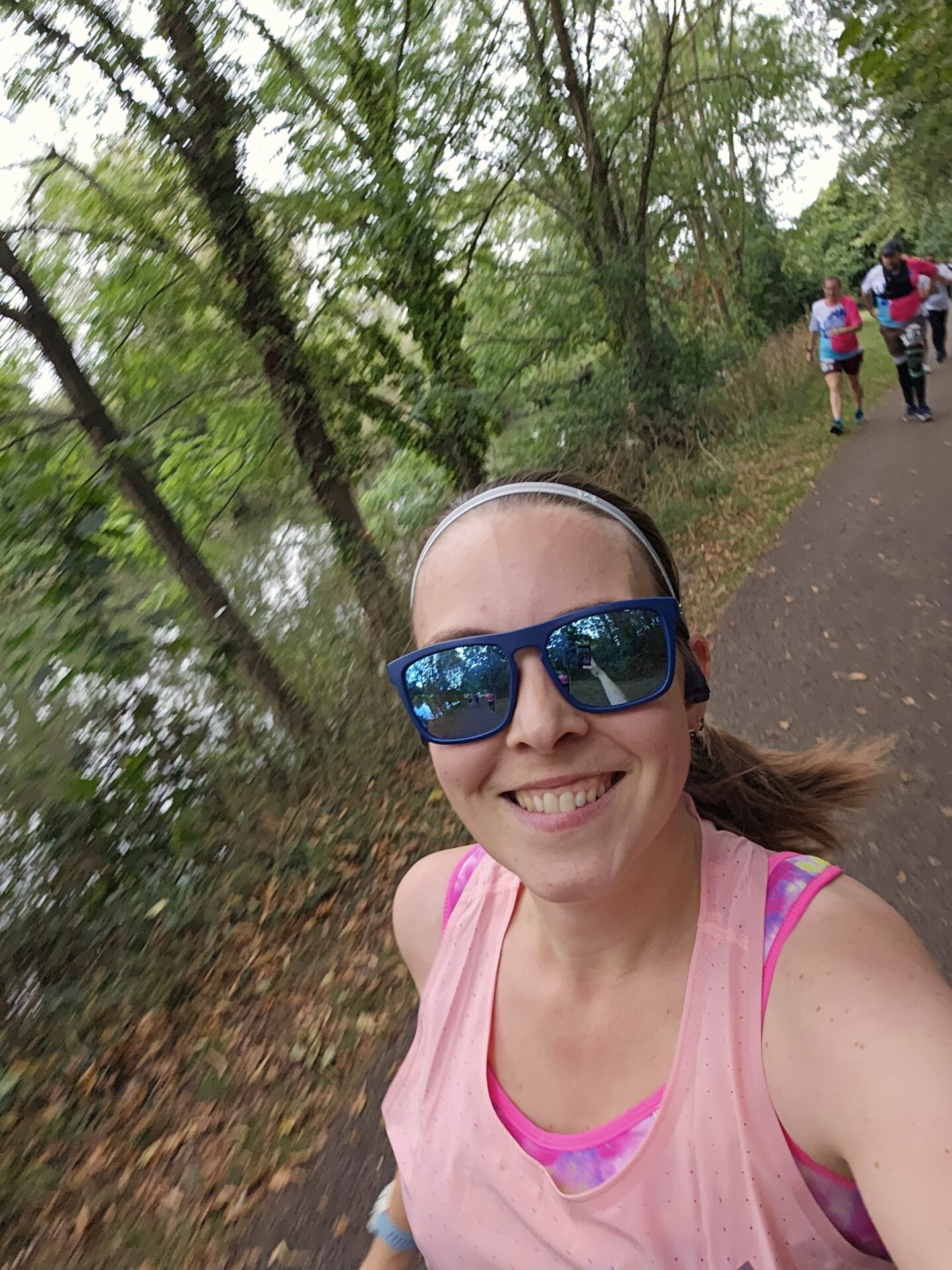 Selfie of a woman in pink tank top and sunglasses as she runs along a footpath