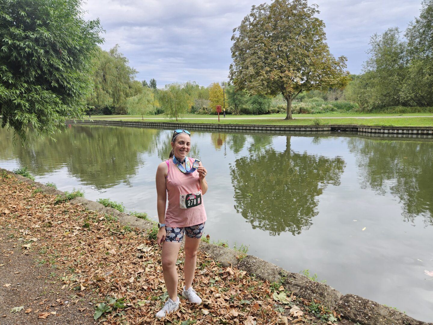 Woman in running kit comprising a pink tank top and shorts is standing in front of the river, holding a medal. She is smiling.