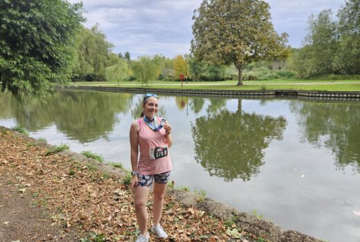 Woman in running kit comprising a pink tank top and shorts is standing in front of the river, holding a medal. She is smiling.