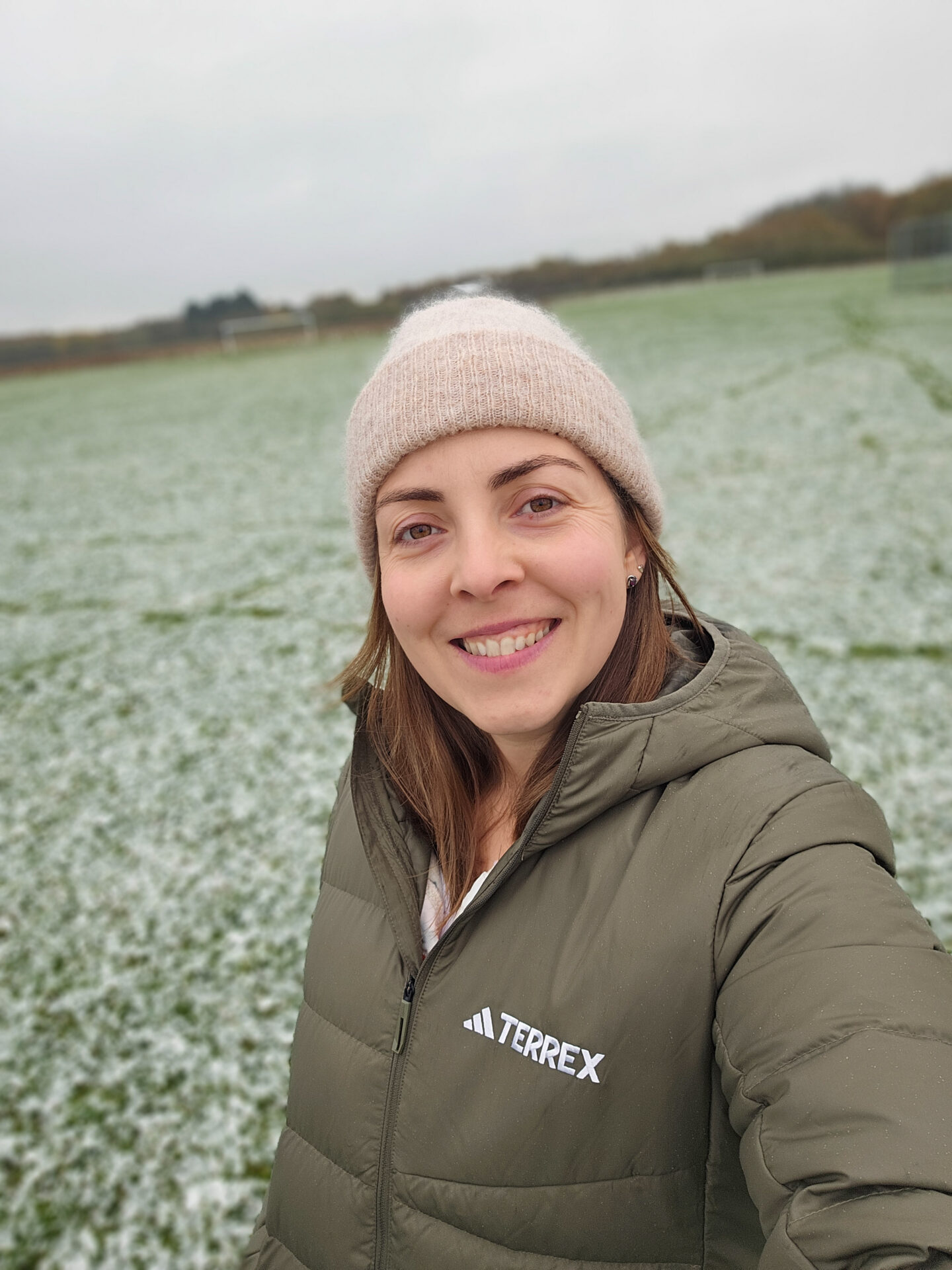 woman wearing a green jacket and beige beanie is smiling at the camera. she is standing in a field full of snow. 