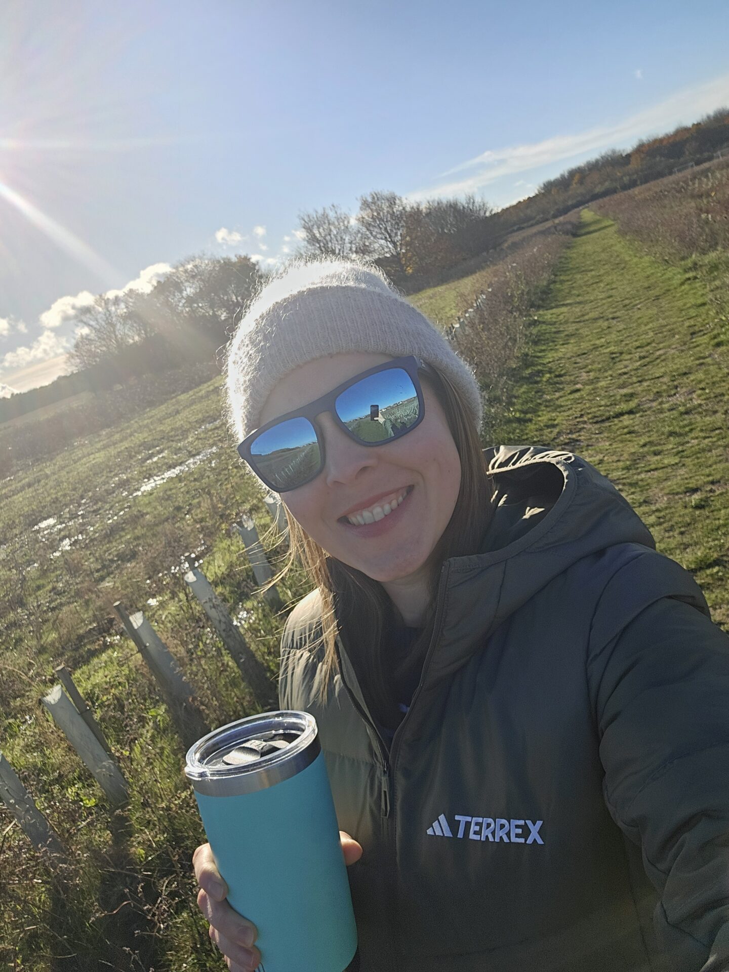 woman wearing green jacket, beige beanie and sunglasses is smiling at the camera. she is holding a green thermos. 