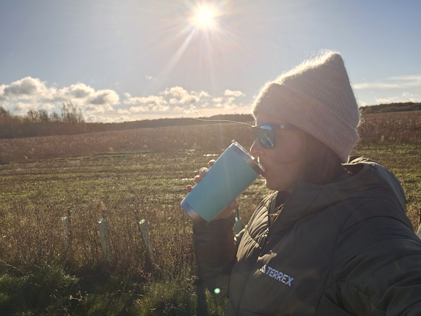 Side profile of a woman in a beige beanie, green down jacket and sunglasses. She is drinking from a green thermos flask. There is a field behind her and bright sunshine in the sky. 