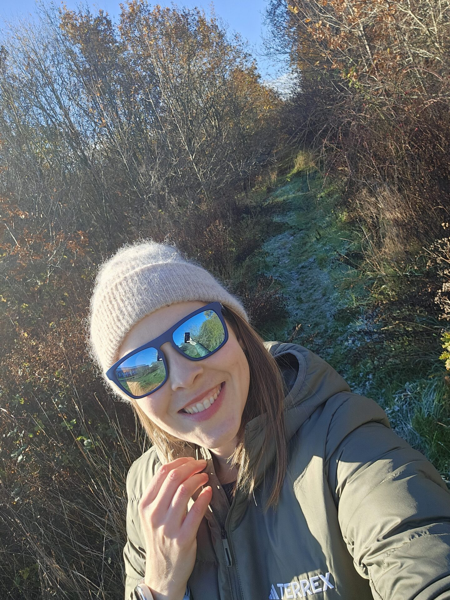 woman wearing a beige beanie and sunglasses is smiling at the camera. A path through the woods disappears behind her. 