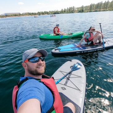 family photo with a man on a paddleboard, girl on a paddleboard and woman sitting in a green kayak. the water is blue with a row of trees in the background