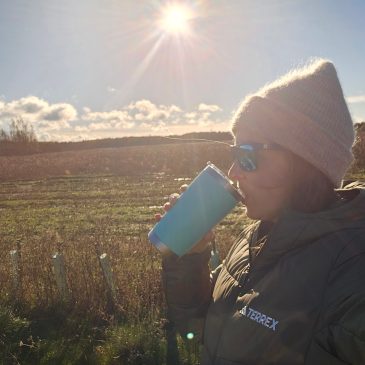 Side profile of a woman in a beige beanie, green down jacket and sunglasses. She is drinking from a green thermos flask. There is a field behind her and bright sunshine in the sky.