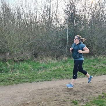 Woman in black leggings, blue running top and green gilet runs along a country trail