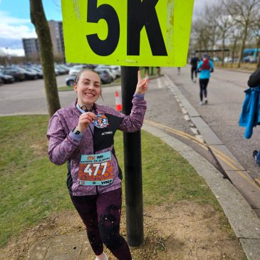 woman in running kit with a medal round her neck, pointing at a large neon yellow sign which says 5k in black