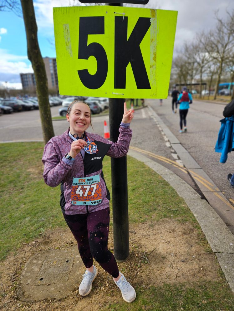woman in running kit with a medal round her neck, pointing at a large neon yellow sign which says 5k in black