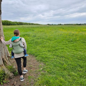 woman carrying baby on her back in carrier. she is leaning on a tree in the middle of a green field.