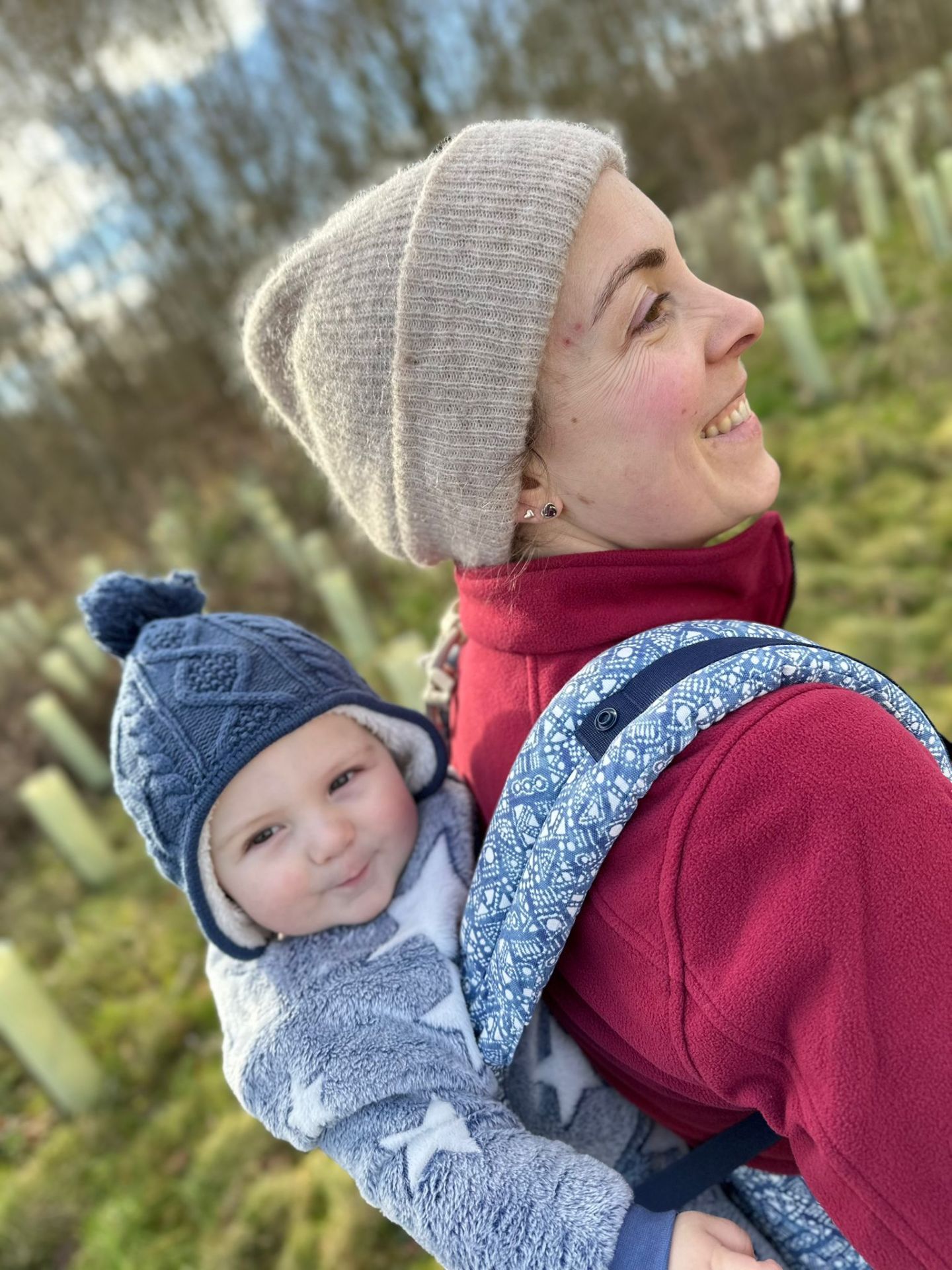 smiling woman in a beanie hat is carrying a toddler in a carrier on her back.