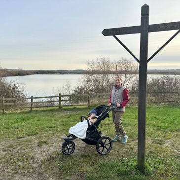 Woman with a running buggy stands on the grass by a wooden sign, with a lake in the background
