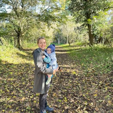 woman in green coat is carrying a baby in a sling. she is smiling at the camera and the baby is wearing a blue woolly hat. They are on a muddy path through the woods with trees behind them.