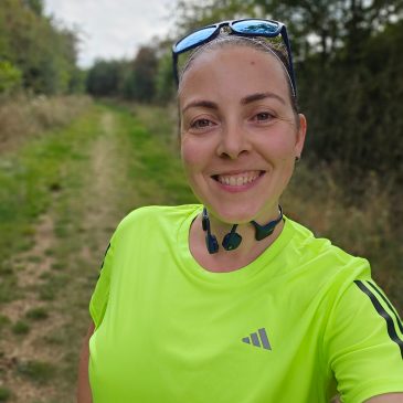 woman in yellow running top smiles at the camera, with a green grassy trail behind her