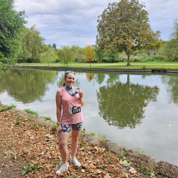 Woman in running kit comprising a pink tank top and shorts is standing in front of the river, holding a medal. She is smiling.