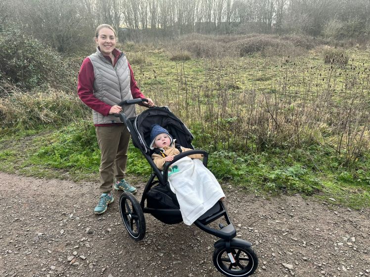 Woman in a red fleece and grey gilet is standing with her hands holding the handlebars of a running buggy with a little baby wrapped up warm inside. Behind them is grass, and trees.