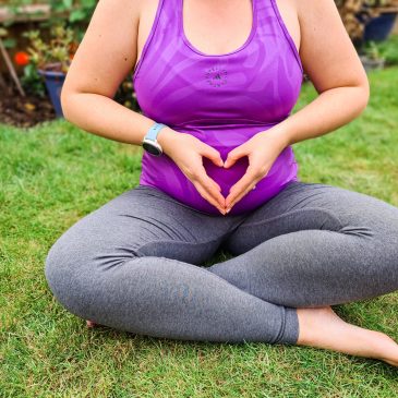 Pregnant women sits cross-legged on the grass, with her hands making a heart on her belly. She wears grey leggings and a purple top.